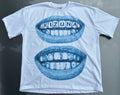 A white short-sleeve t-shirt laid flat on a dark gray surface, featuring a blue graphic of two open mouths with teeth, where the top mouth has the word 'KIZUNA' spelled out in letter tiles.