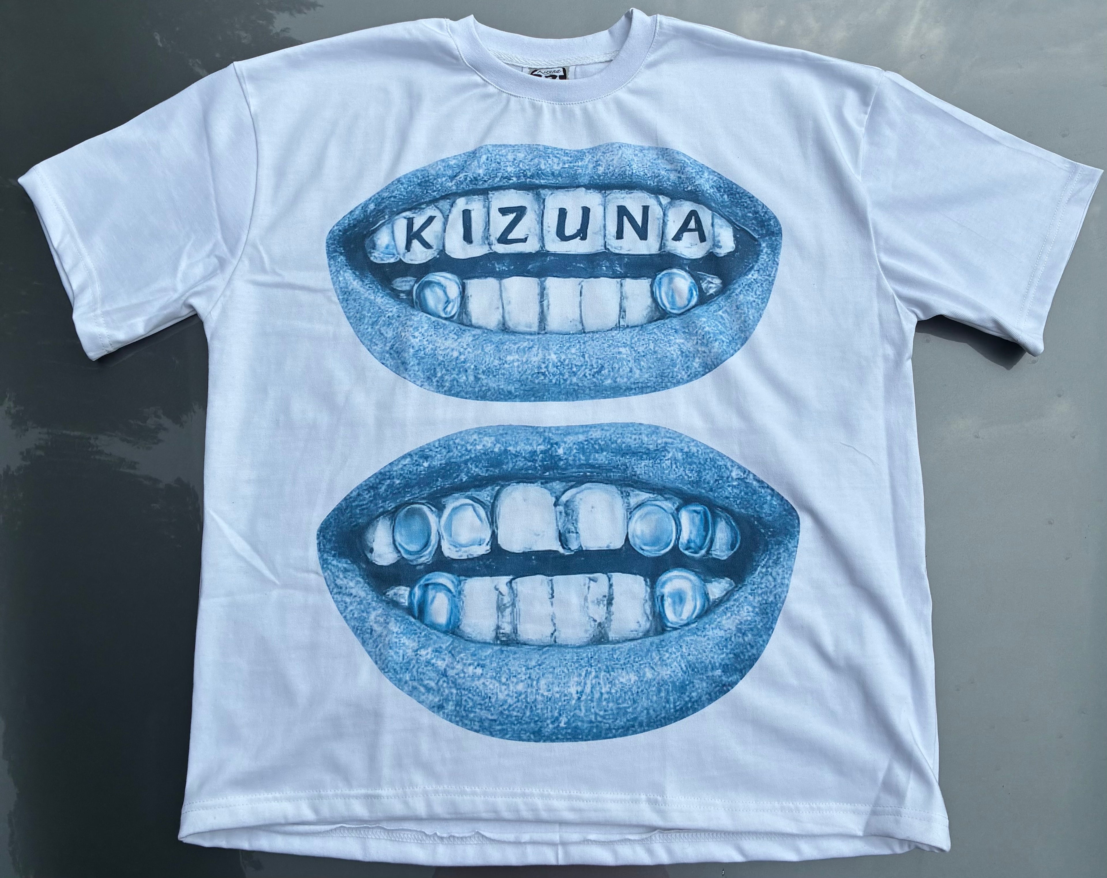 A white short-sleeve t-shirt laid flat on a dark gray surface, featuring a blue graphic of two open mouths with teeth, where the top mouth has the word 'KIZUNA' spelled out in letter tiles.