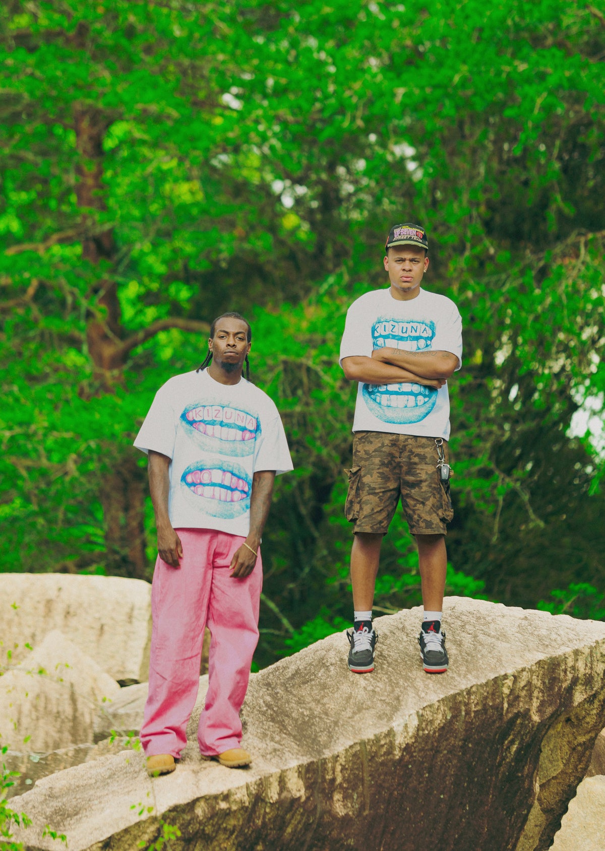 Two men standing on large rocks in a lush green outdoor setting, wearing matching white graphic t-shirts with blue and pink designs, one paired with pink sweatpants and the other with camouflage shorts.